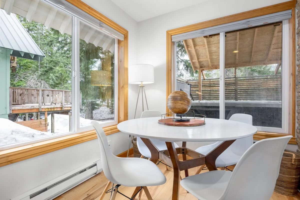 A bright, inviting dining area with a round table, large windows flooding the space with natural light, and comfortable seating. The backdrop suggests a cozy, family-friendly atmosphere ideal for group meals, games, or conversations while enjoying views of the surrounding neighborhood.