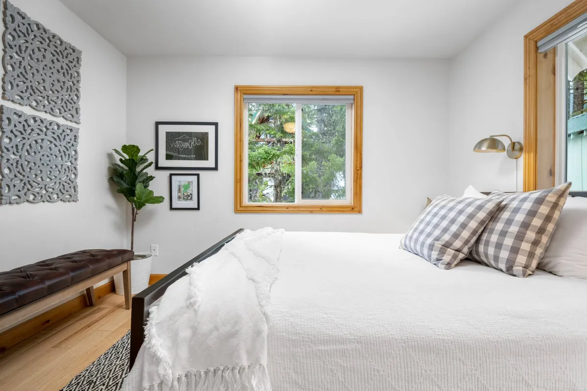 A tranquil king bedroom with white bedding, checkered pillows, and a large window that lets in natural light. The room is accented by decorative wall art, a potted plant, and a bench seat, creating a serene retreat for restful moments or waking up to mountain views.