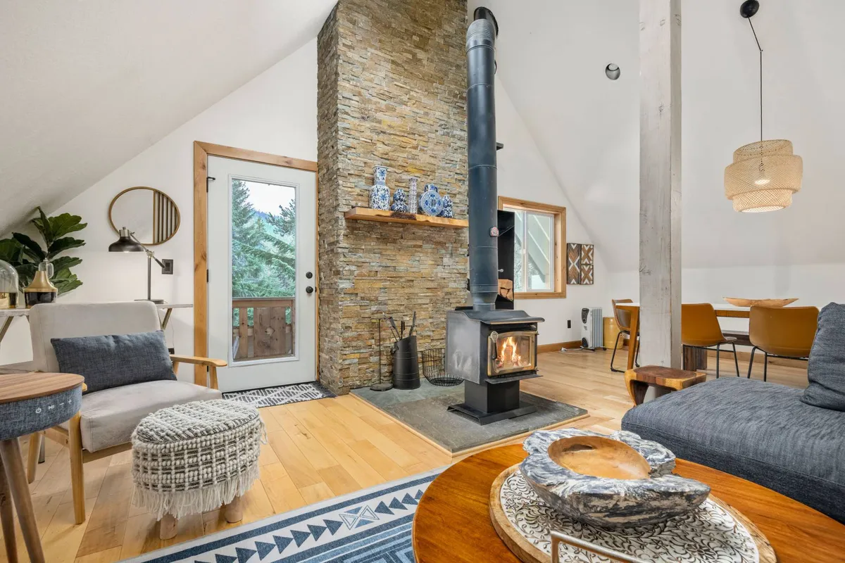 The image showcases a warmly lit, inviting living and dining area with a grand stone fireplace as the centerpiece, flanked by a cozy seating arrangement and a rustic wooden dining table. Soft natural light filters in, highlighting decorative blue ceramics and offering glimpses of the surrounding wilderness. The scene conveys a sense of comfort, togetherness, and mountain charm.