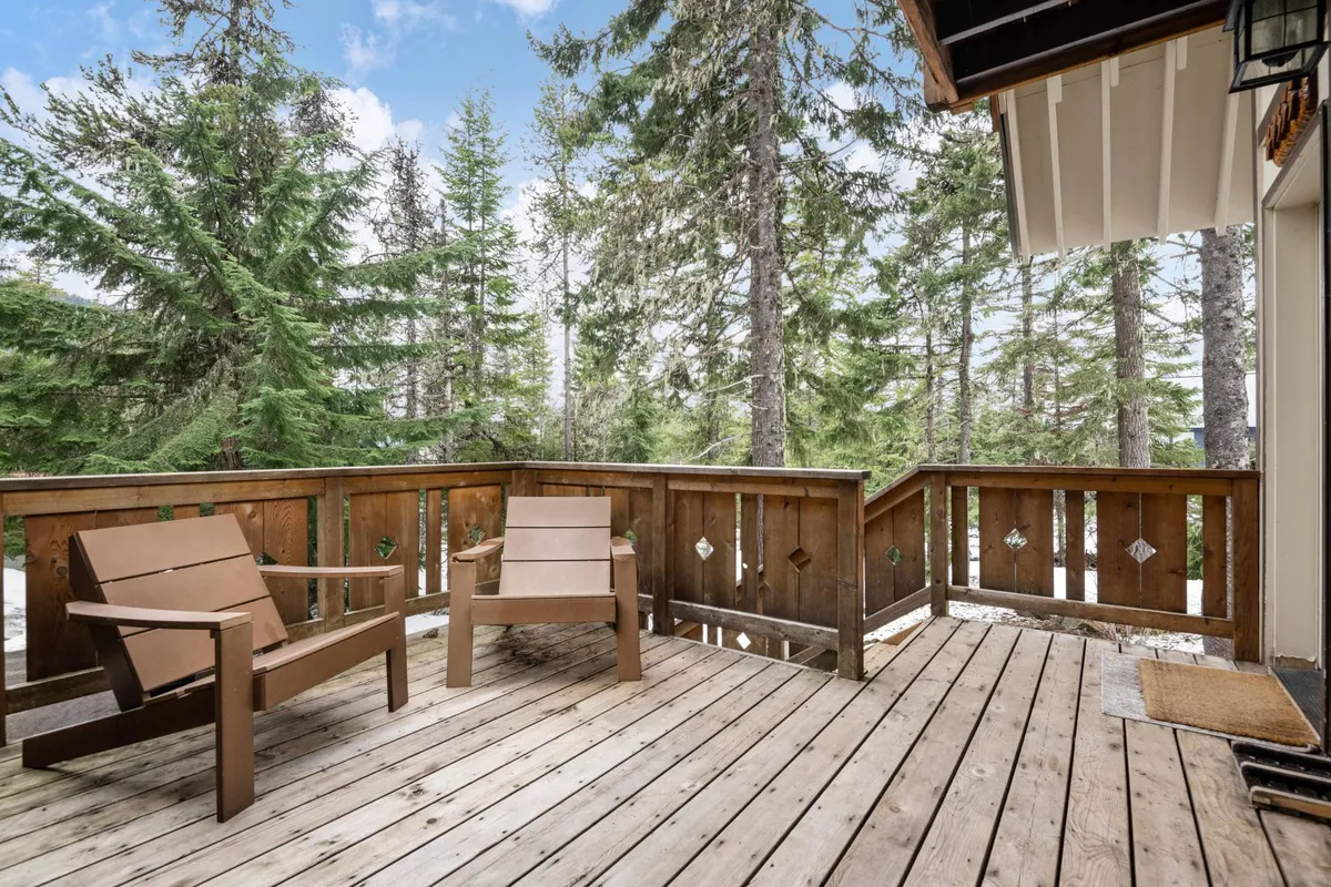 A set of wooden chairs arranged on a deck with a wooden railing, nestled among tall trees. The setting feels tranquil and encourages relaxation, reading, or sharing stories outdoors, with the forest as a backdrop.