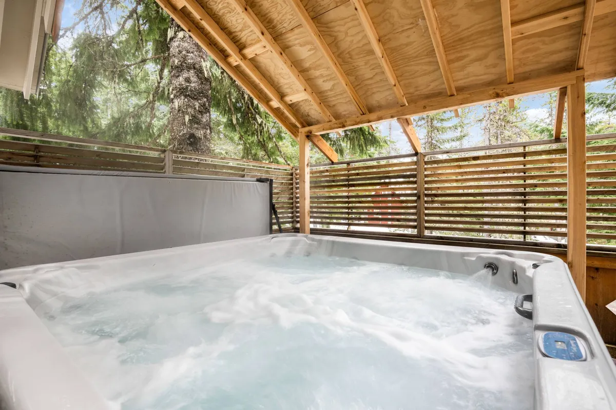 An inviting outdoor hot tub framed by rustic wooden beams and surrounded by lush greenery (which, in winter, can be imagined as snow-covered), offering a relaxing space for après-ski unwinding and socializing with friends or family.