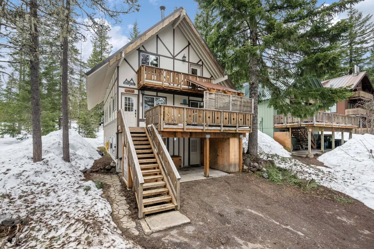 The cozy exterior of Mountain Rose, featuring wooden steps leading up to the entrance, a balcony with a wooden railing, and surrounding snow-laden trees. The image conveys a warm, inviting basecamp atmosphere in a picturesque winter setting.