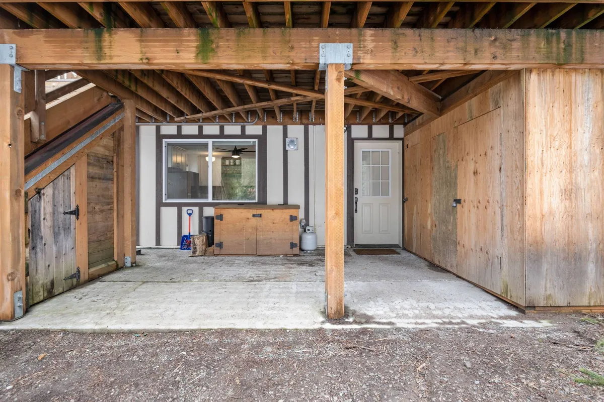 This image presents the exterior entryway of Mountain Rose, featuring welcoming wooden beams and the entrance door. The cozy covered area hints at a sheltered spot for gathering or storing gear, with a storage box visible. The view connects the home to its peaceful, walkable environment, suggesting ease of access for all ages.