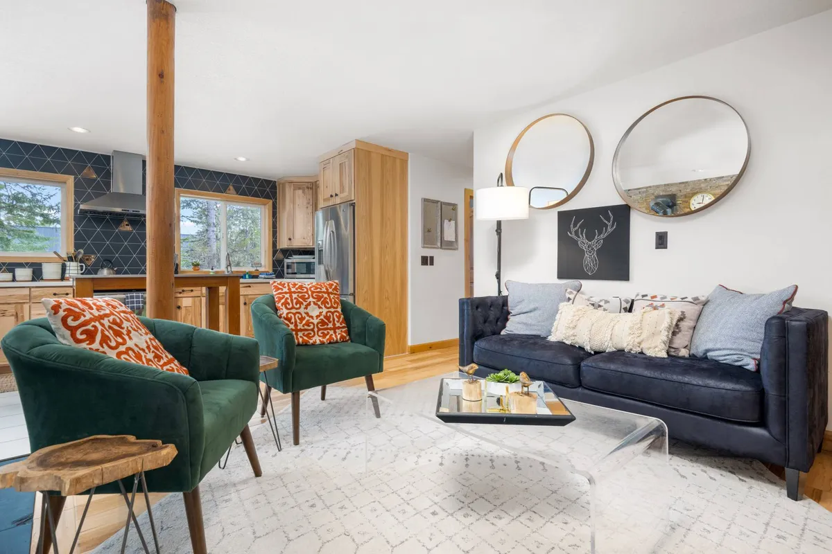 A stylish, open-plan living area features green chairs, a comfortable sofa with decorative pillows, round mirrors, and a visible kitchen area in the background. The space is designed for socializing, entertaining, and seamless transitions between cooking and relaxing.