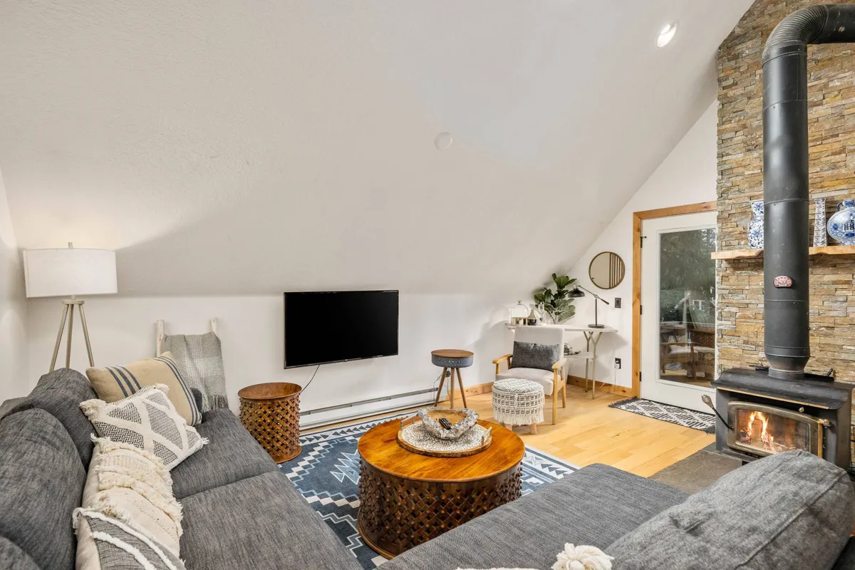 A warm, welcoming living area centered on a rustic stone fireplace, with a comfortable gray sofa and decorative pillows. The ambiance suggests cozy moments spent gathered by the fire after winter adventures.