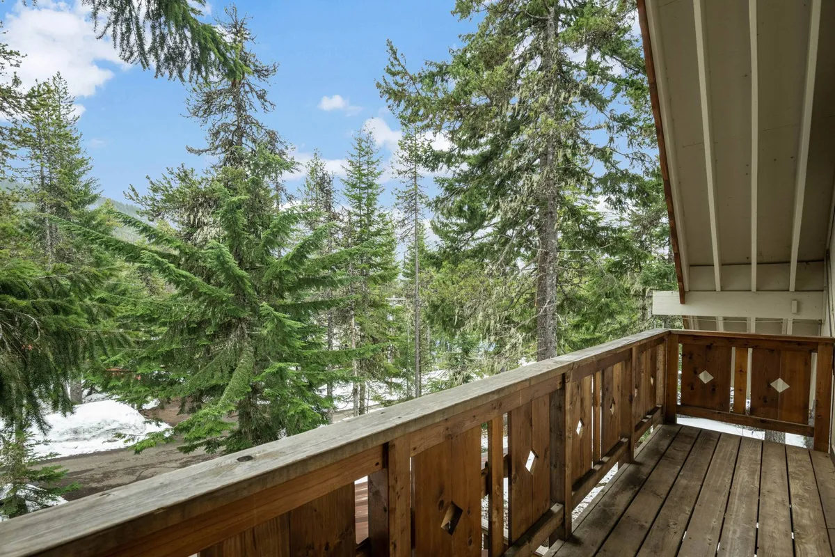 A wooden balcony railing overlooking tall evergreen trees with a bright blue sky above, creating a sense of serene immersion in the forest. The vantage point suggests a relaxing outdoor space for morning coffee or bird watching.