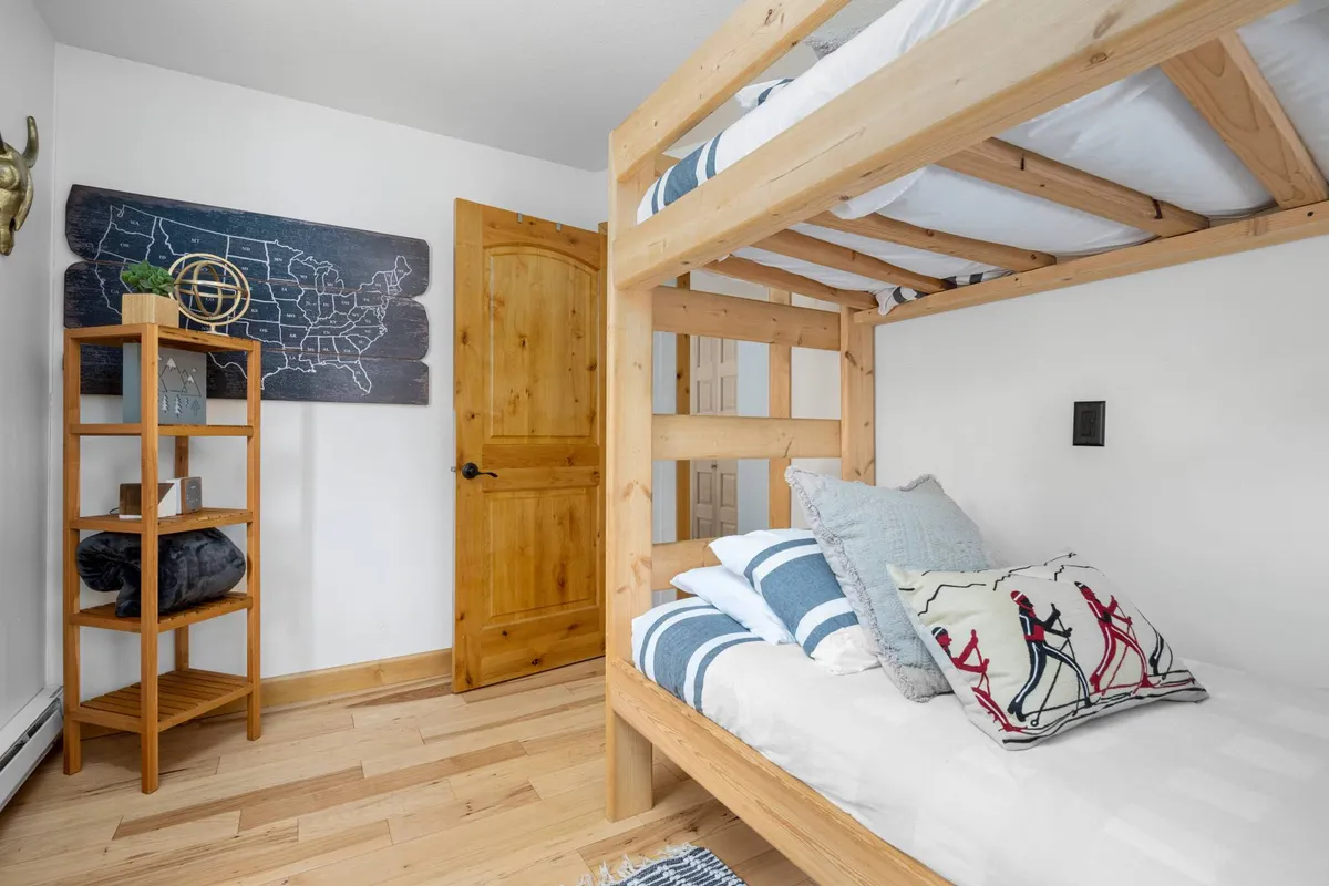 A playful bunk room featuring a sturdy bunk bed, charming wooden shelving filled with books and toys, a colorful pillow, and a wall map for adventure and fun. The space feels cozy, inviting, and ideal for kids or young-at-heart guests.