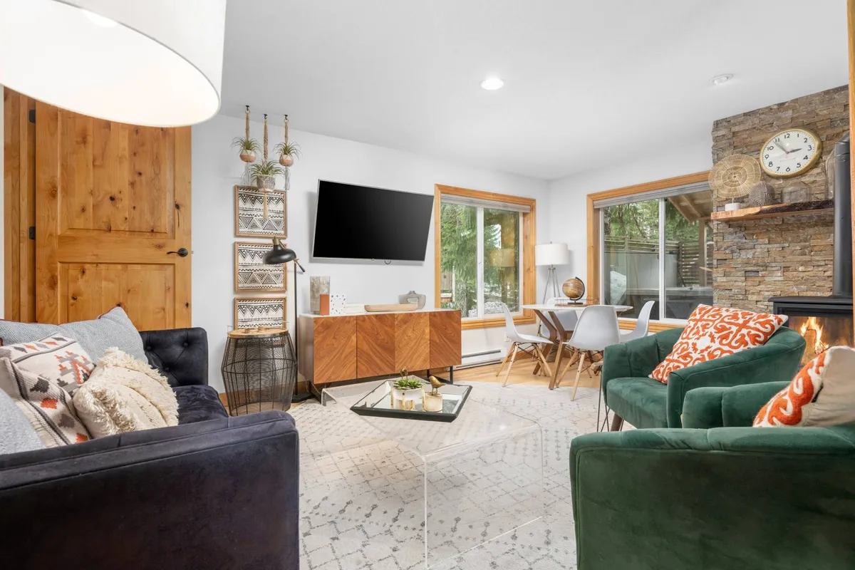 A cozy interior shot featuring plush green velvet chairs, a wooden side table, and a decorative wall clock. Large windows let in abundant natural light, giving the space a welcoming atmosphere perfect for relaxing with loved ones—including pets.